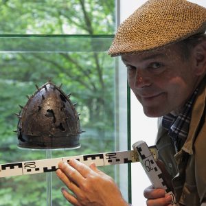 Pago Balke mit einer Mütze und einem Maßstock vor einem altem Helm.